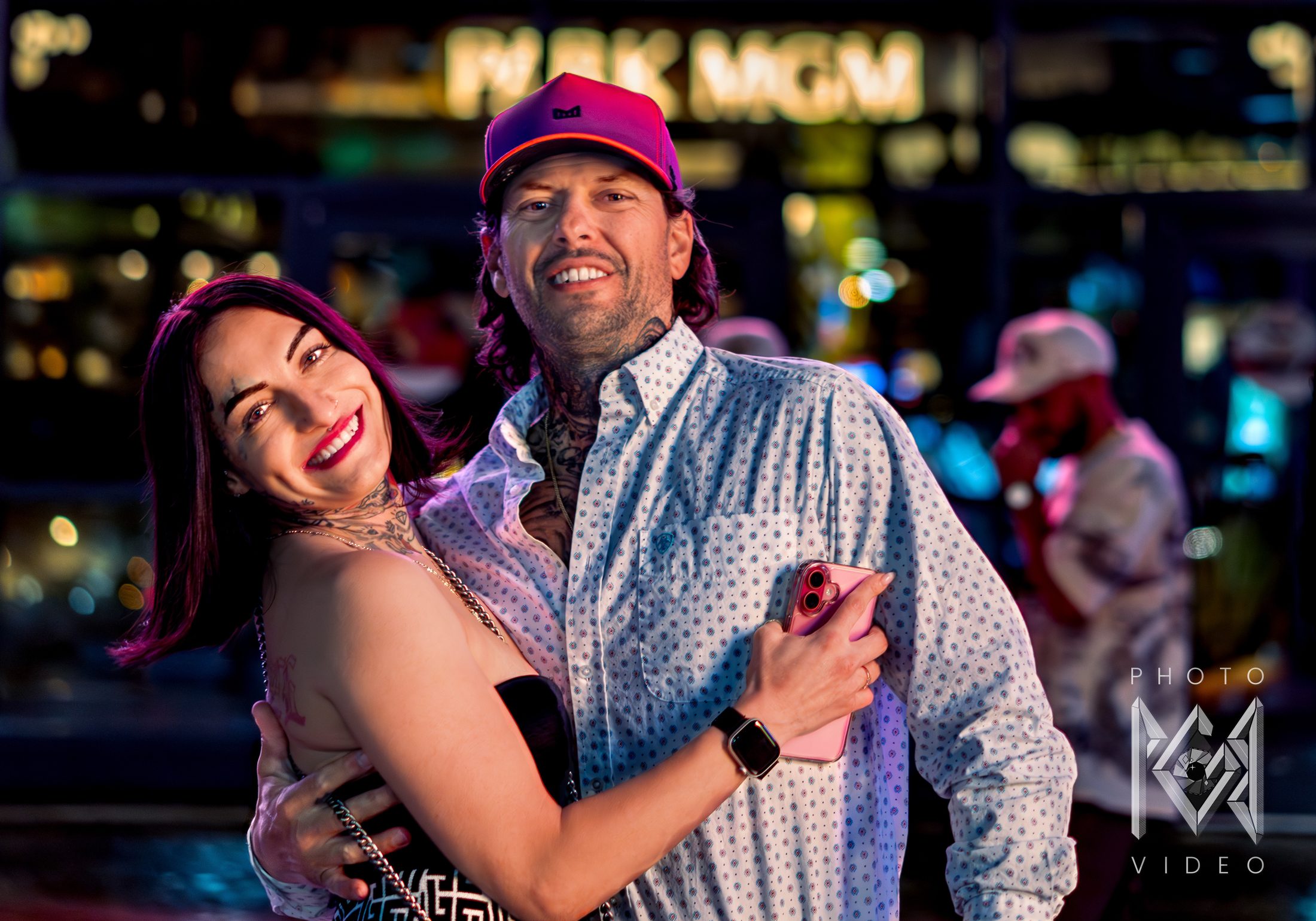 A smiling couple embracing at night, she in red lipstick and he in a pink cap and patterned shirt, with blurred casino lights behind