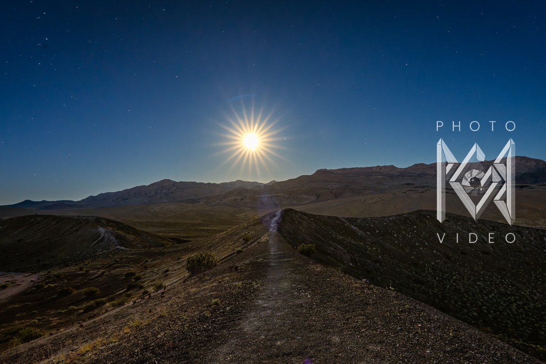 A starburst moon low over a long desert ridge, a path leading toward it under a deep blue dusk sky