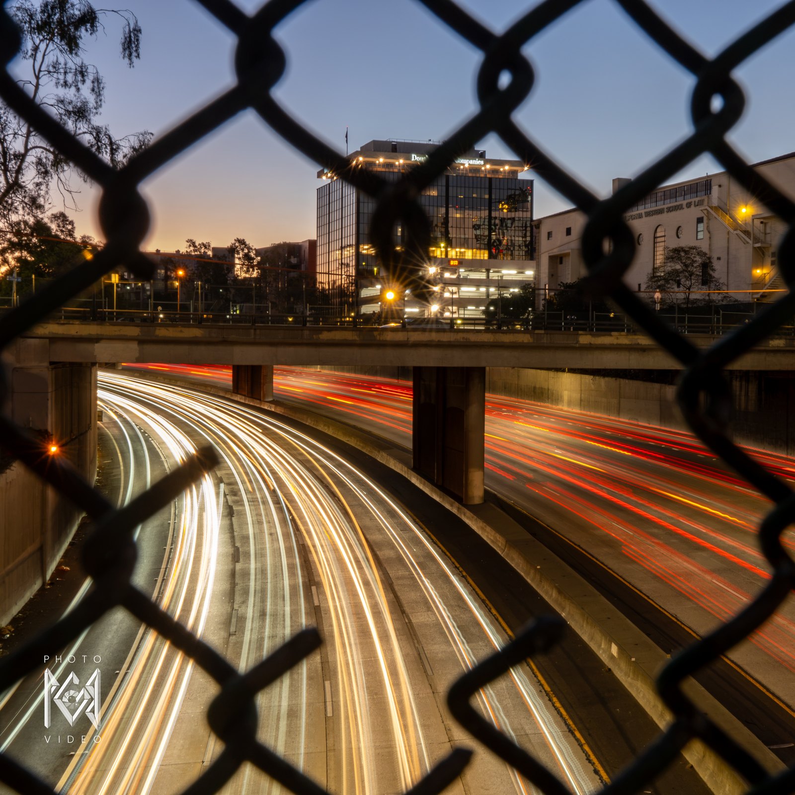 Streaks of white and red car lights curving along a sunken freeway at dusk, seen through a chain-link fence with downtown buildings beyond